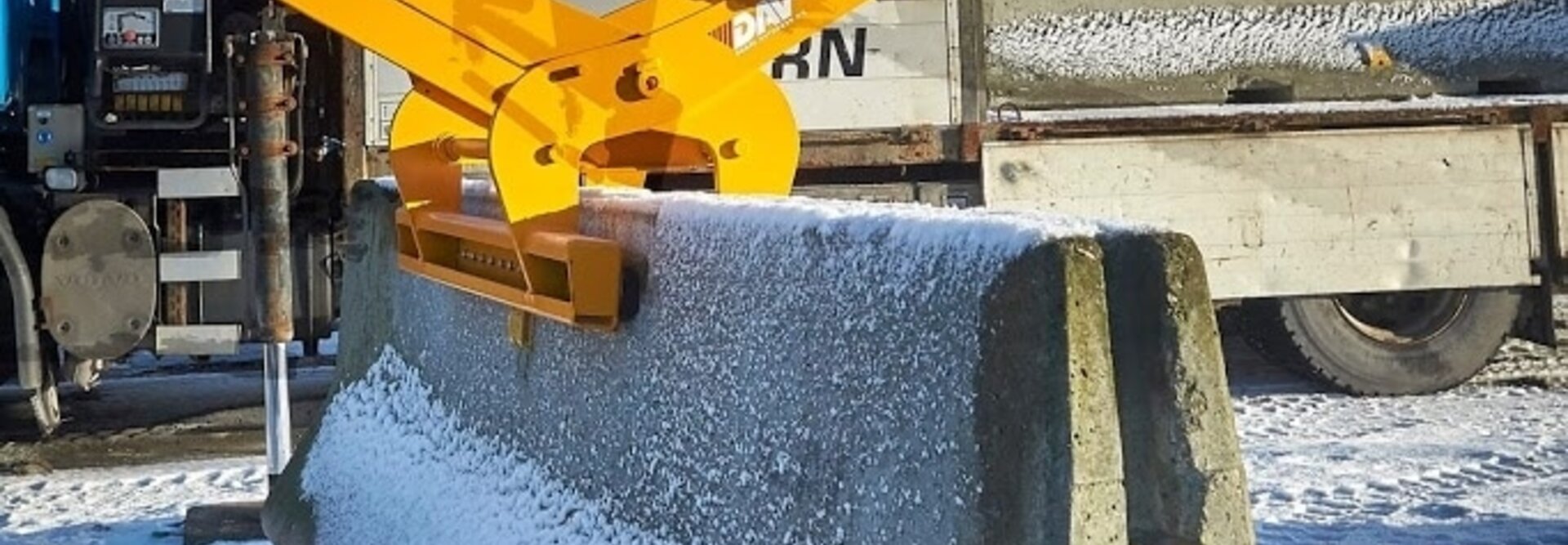 An image of a guardrail lifting clamp from BSV lifting a large concrete block seen from the side. There is snow on the ground and around the construction site, and the concrete block is securely suspended in the air.